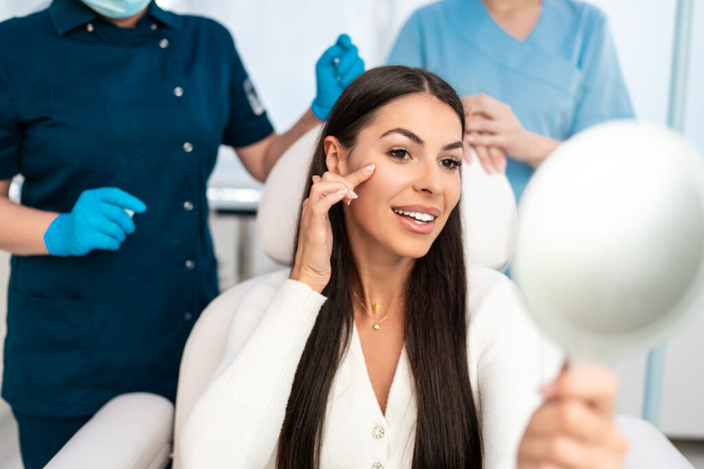 Woman in a skincare clinic examining her skin in a mirror, with two professionals in the background, illustrating the treatment experience for chemical peels and microneedling.